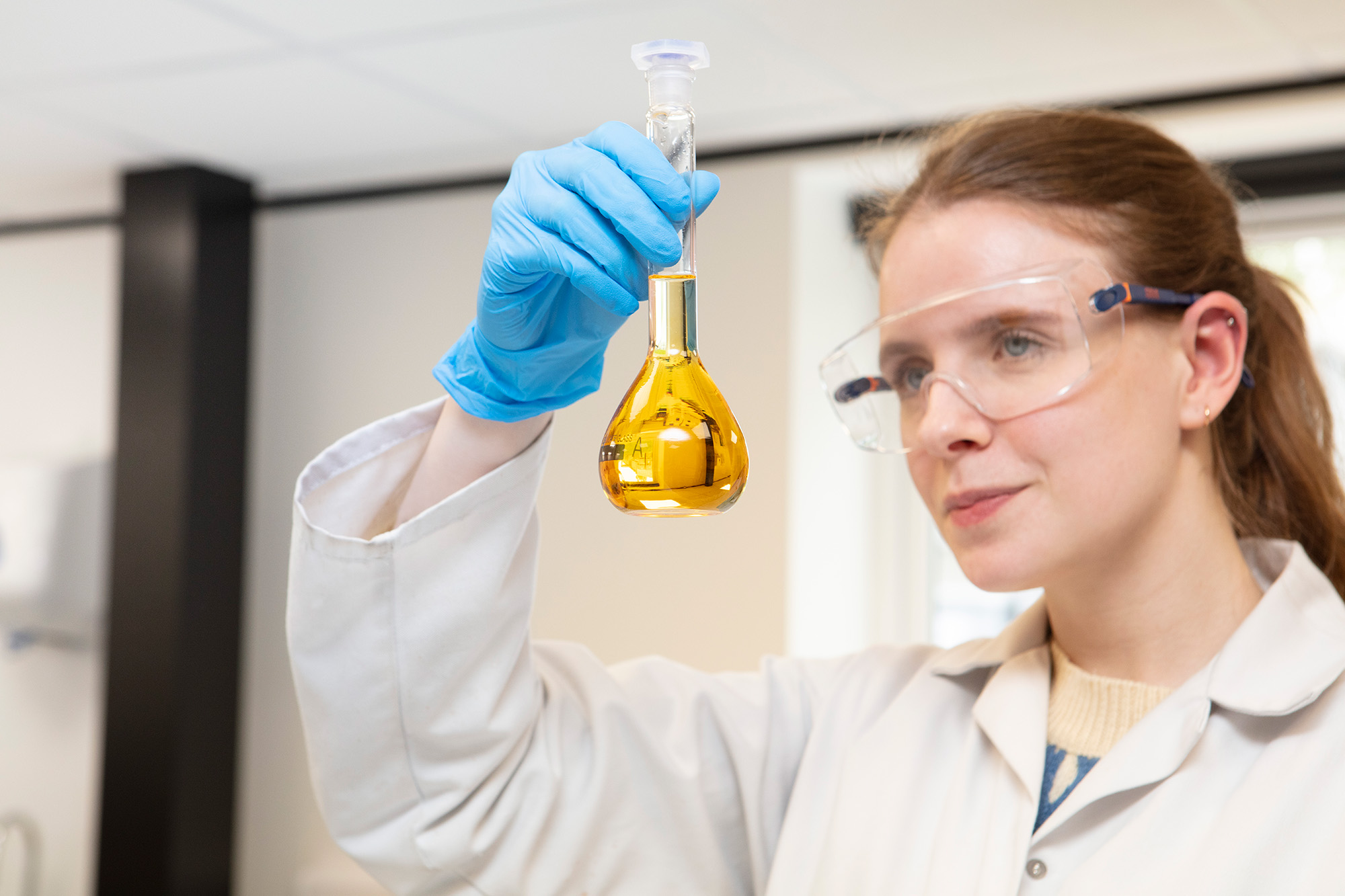 Female Scientist wearing lab coat holding glass vile containing clear yellow orange liquid, a reflect of the room can be seen in the glass.