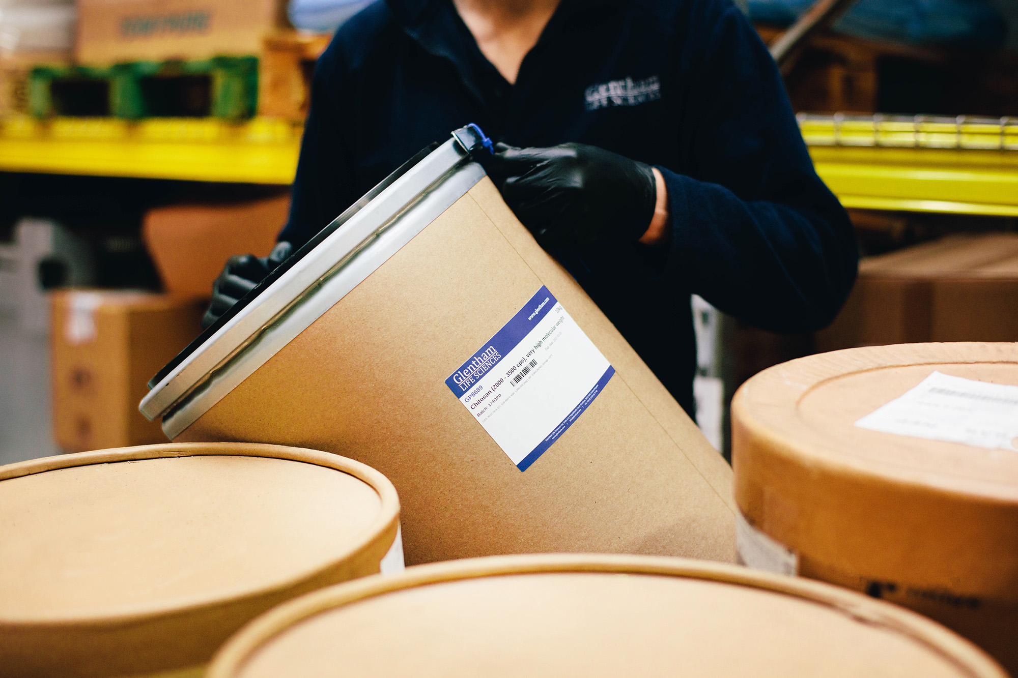 Photo of male warehouse worker lifting a 10kg drum containing Chitosan, the worker is wearing black gloves and a dark blue jumper. He has 3 other drums next to him.